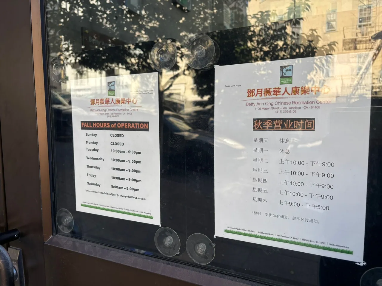 Paper schedules for the Betty Ann Ong Chinese Recreation Center are taped to a window. The city street and trees are reflected in the glass.