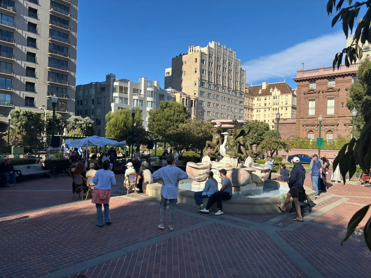 An audience is gathered near a foundation in a brick-lined public square on a clear blue-skied day. Musicians are performing in front of them in a shady area. Buildings loom in the background.