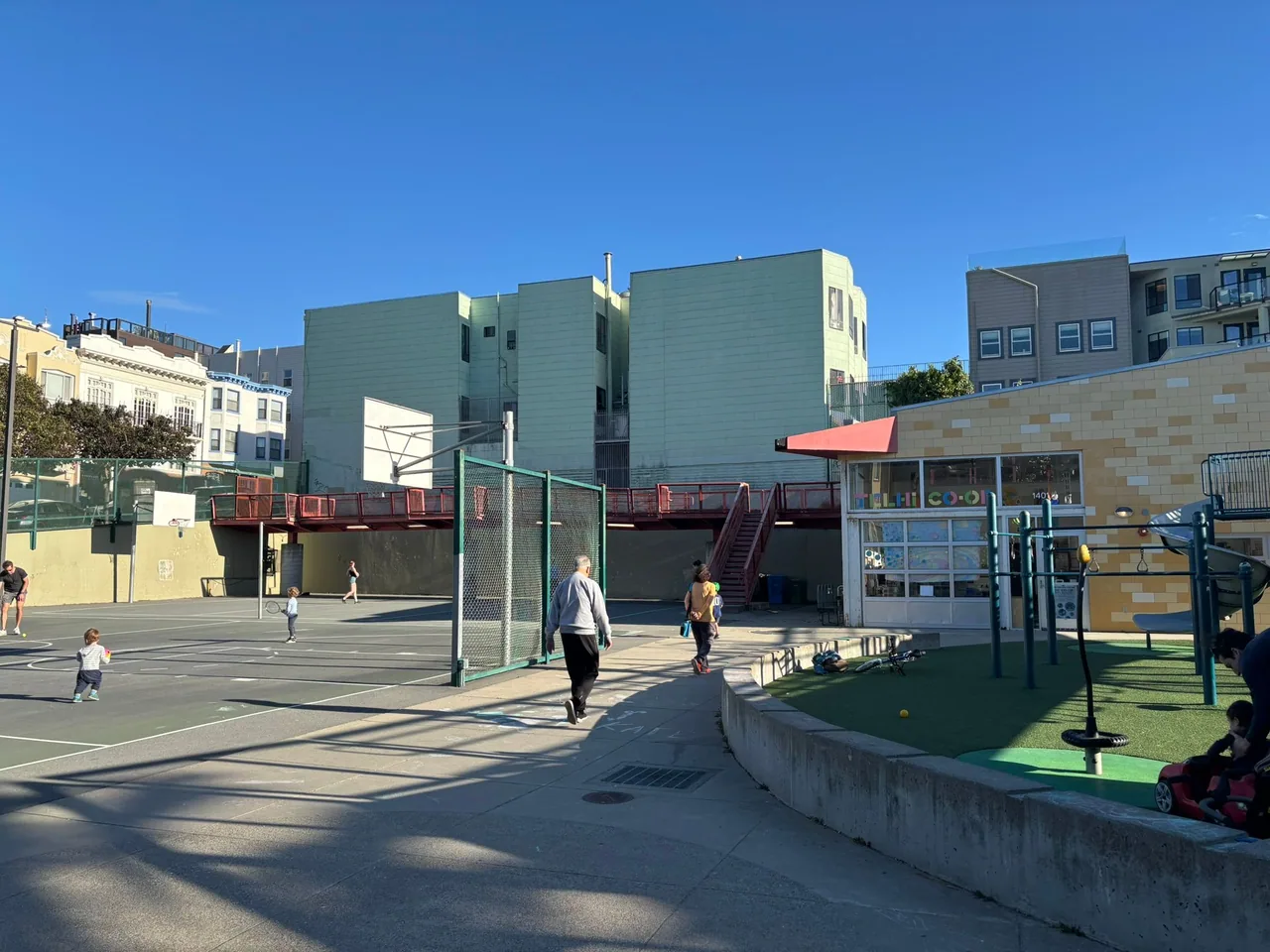 Children and adults play on a large basketball court and playground area. Residential buildings and a clear blue sky are situated in the background.