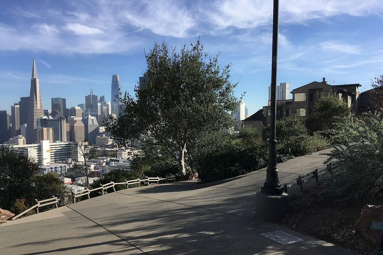 A paved hillside path with railings and plantings overlooks the downtown San Francisco skyline from Ina Coolbrith Park.