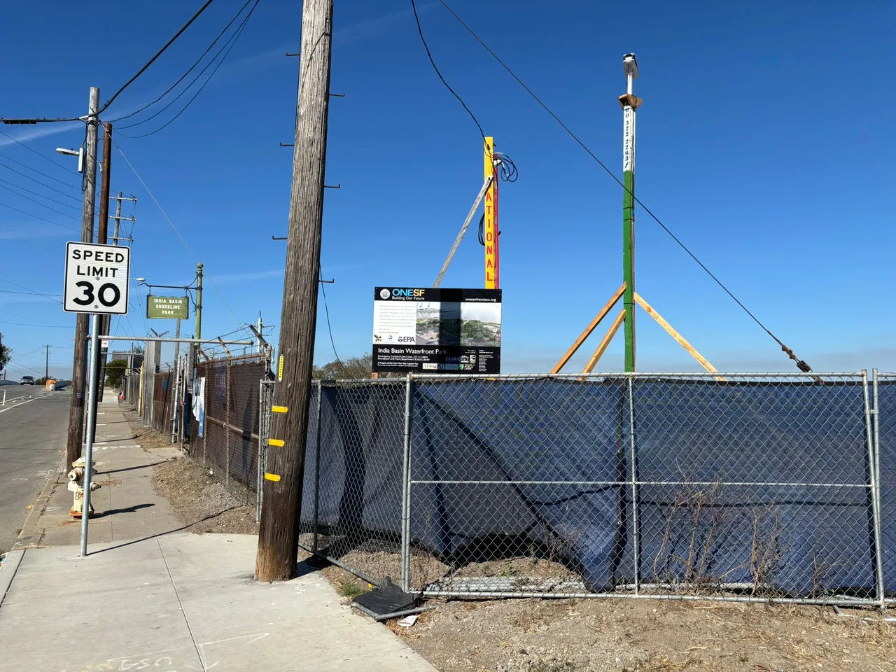 Fenced waterfront construction site beside a narrow sidewalk and utility poles