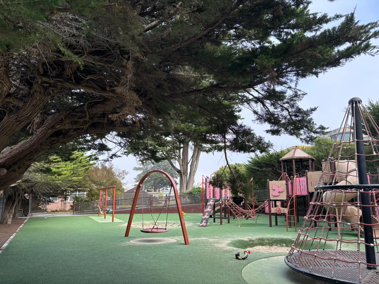 A large tree hangs over an empty playground. A variety of play structures are situated in an an artificial turf playing area.