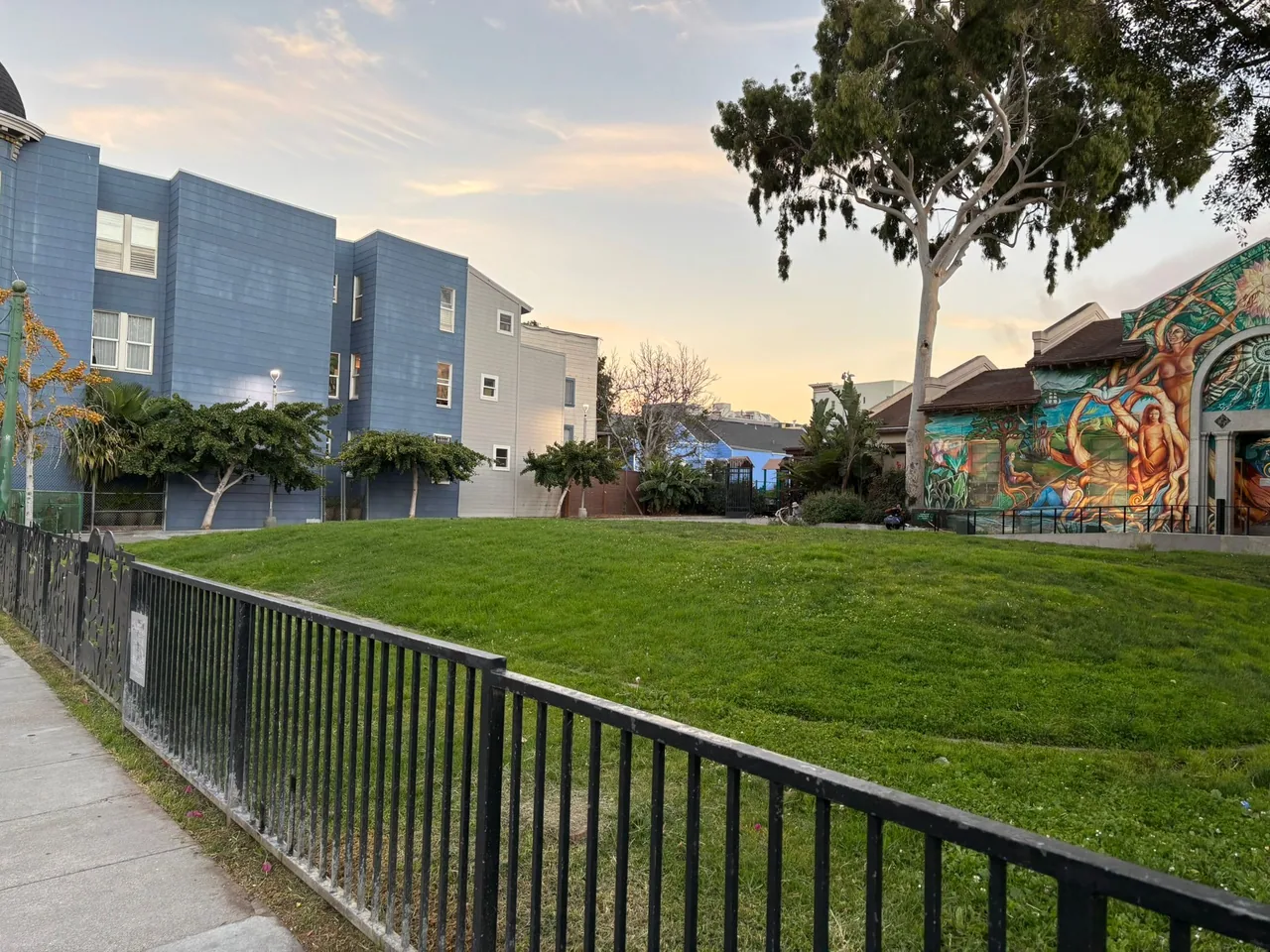 A colorful mural adorns a park building near a flat patch of green grass. A black metal gate in the foreground separates the sidewalk from the grass. Residential buildings, trees, and a sunset sky can be seen in the background.