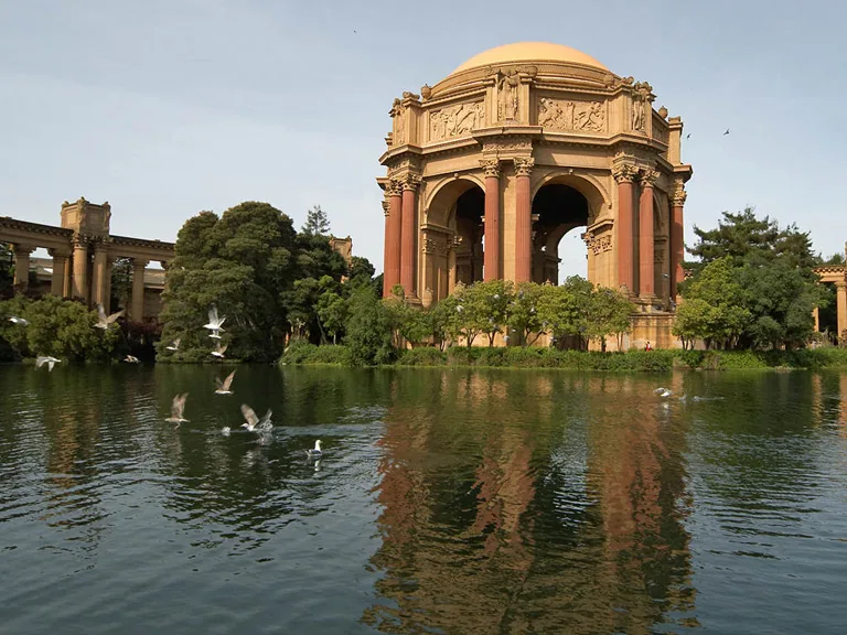 The Palace of Fine Arts rotunda rises behind trees and a lagoon with birds flying above the water.