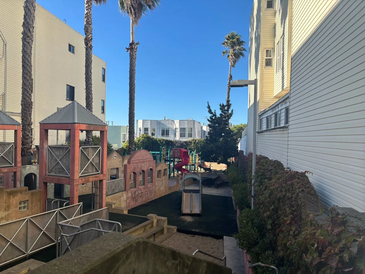 A mini park filled with playground structures sits empty on a blue-skied day. The park is surrounded on two sides by city buildings. Several palm trees decorate the park.