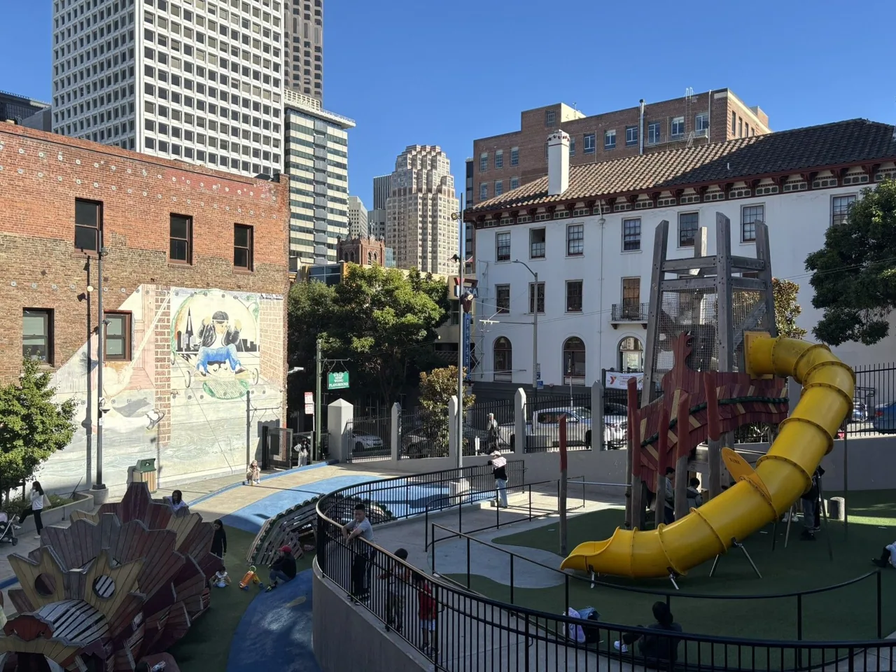 Children play in an urban playground on a clear blue day. Major features are a large structure with a curved yellow slide and a dragon-shaped play structure.